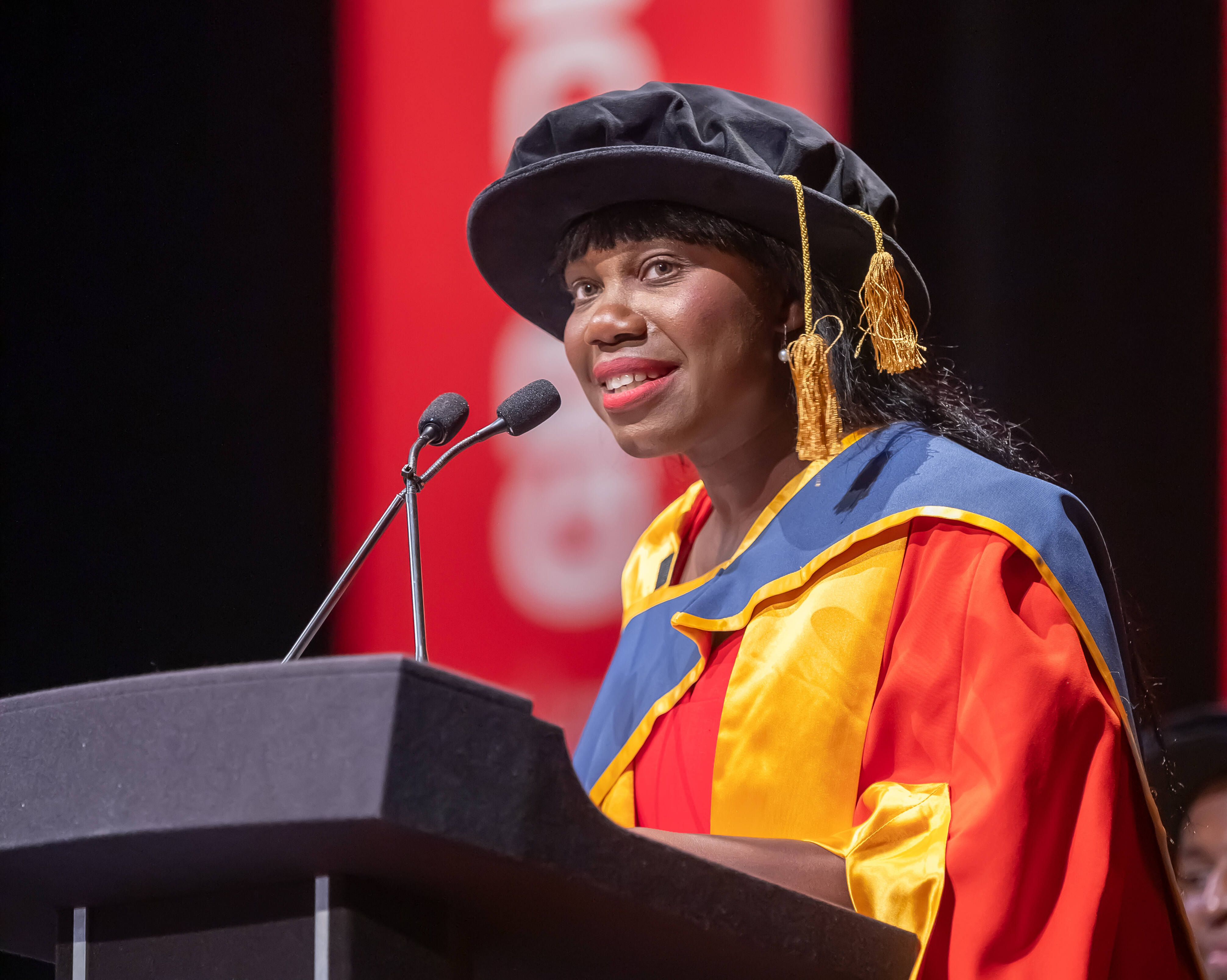 Dr Sally Penni giving a speech in red, yellow and blue graduation attire on stage in front of a large red banner. 